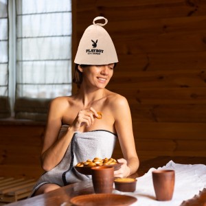 Young woman in the sauna with a cap on her head sits at a table with round bagels, honey and tea enjoying a wellness day
